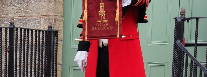 Town crier dressed in a red and black ceremonial outfit holding a decorative banner, standing on steps beside a handbell in front of large green doors.