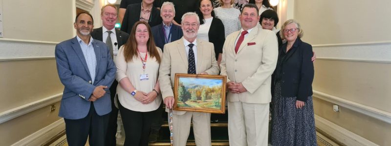 Group photo on an ornate staircase landing, featuring several people standing together while one person at the front holds a framed landscape painting.