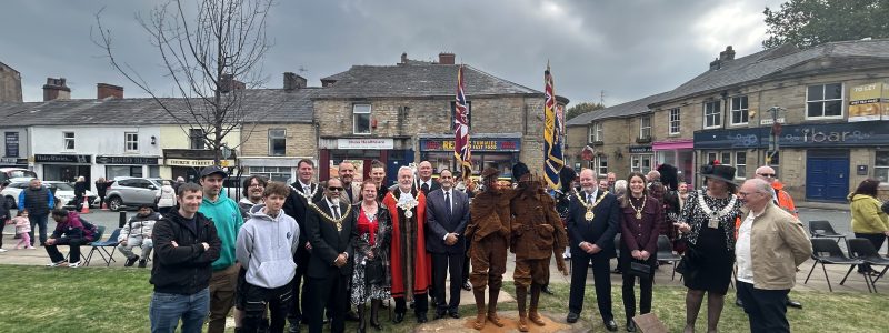 Group gathered at an outdoor ceremony with uniformed participants, civic representatives, flags and a central drum marked “Accrington Pipe Band,” set in a town square with stone buildings in the background.