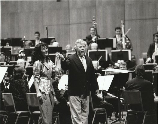 Orchestra performance scene with two people standing at the front of the stage, surrounded by seated musicians and music stands.