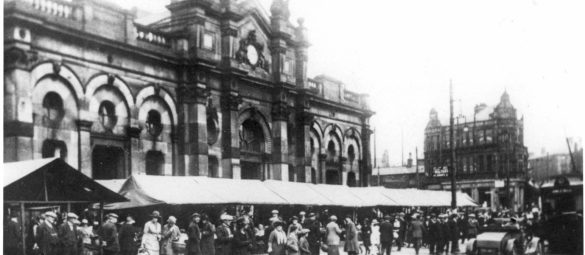 Crowded outdoor market scene outside a large arched stone building, with rows of covered stalls and many people gathered along the street.