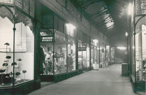 Historic indoor shopping arcade with illuminated shopfronts, displaying clothing, hats and various goods beneath a high arched glass roof.