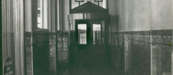 Historic tiled corridor leading to a wooden revolving-style entrance with glass panels and signage visible on the doors.