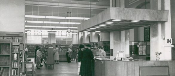 Historic library interior showing a large central counter, tall bookshelves, and visitors browsing and speaking with staff.