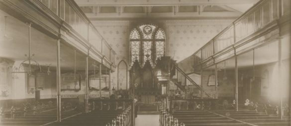 Interior view of a historic church showing wooden pews, upper galleries, decorative wall patterns and a large stained‑glass window above the altar.
