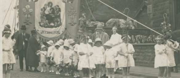 Historic parade scene featuring a group of children in light‑coloured outfits holding decorated staffs and flowers, gathered in front of a large “Parish Church of St. James Accrington” banner on a cobbled street.
