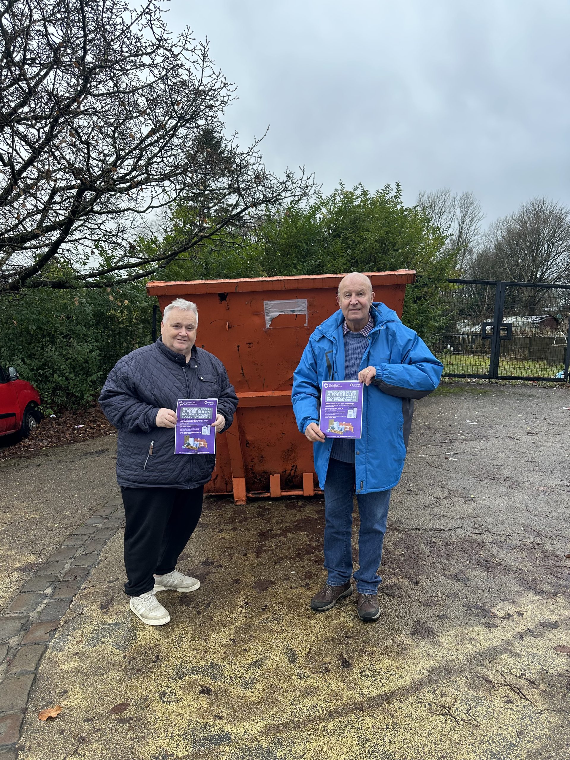 Two Councillors holding bulky waste collection leaflets infront of large, orange skip