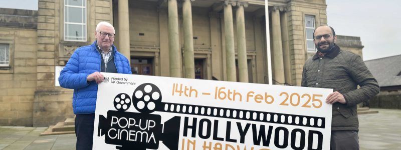 Two men holding Hollywood in Harwood sign. Mercer Hall building and flagpole in background