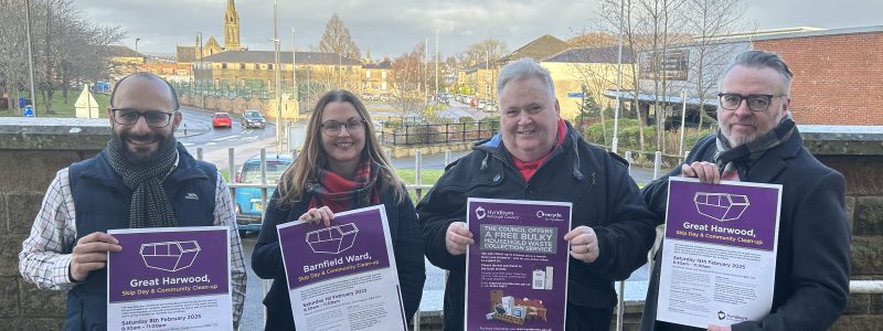 Four Councillors holding skip day posters. Buildings, blue sky and clouds in background