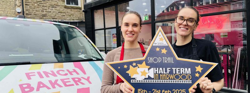 Two people standing outside Finch Bakery, holding a star-shaped sign promoting a local shop trail from February 15th to 21st, 2025. A branded van is parked nearby.