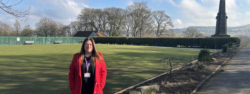 A person standing on a step in front of a bowling green. Large War memorial in background.