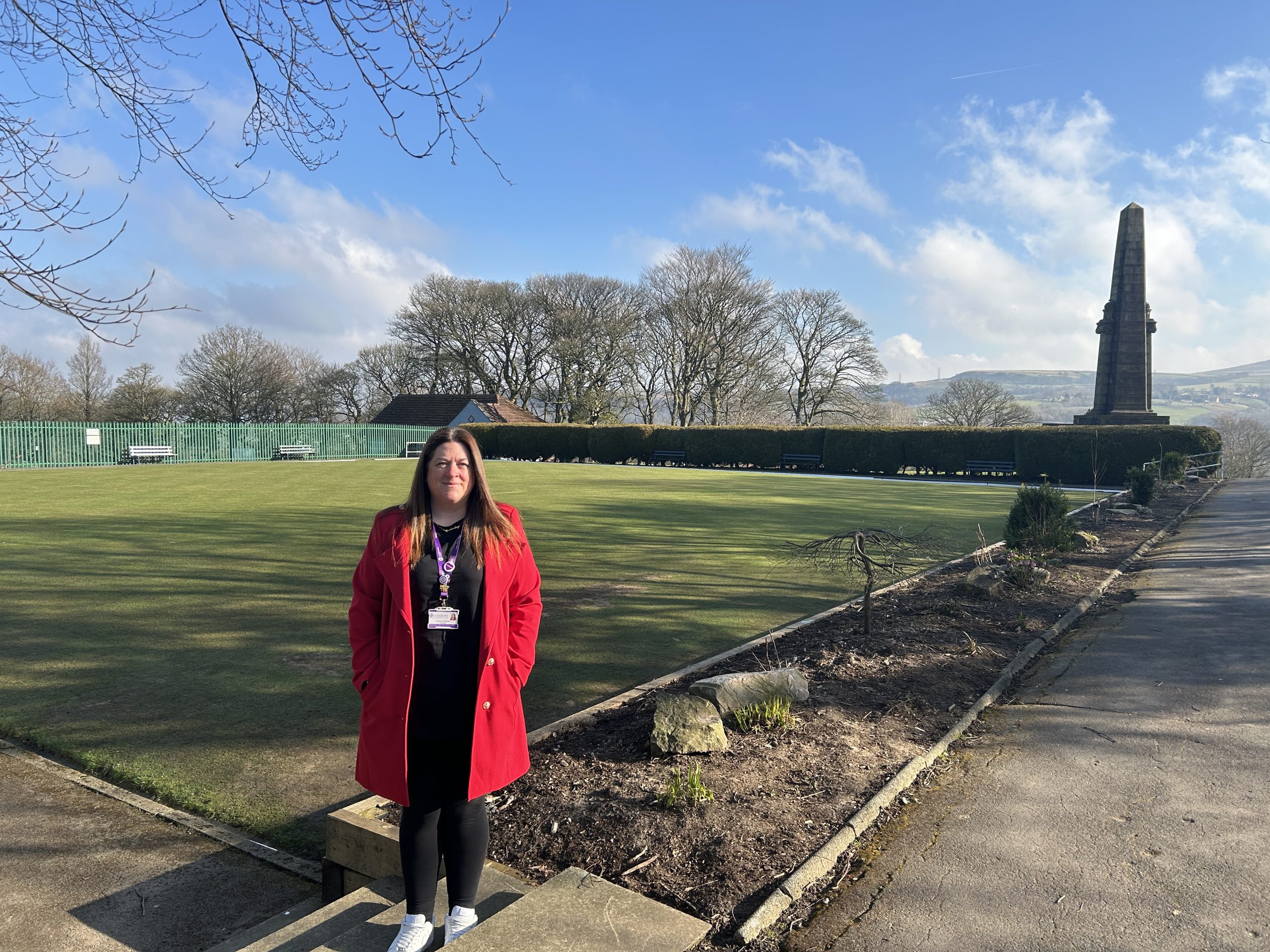A person standing on a step in front of a bowling green. Large War memorial in background.
